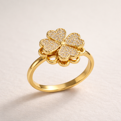 A gold ring featuring a four-petal flower design with heart-shaped centers paved with small clear stones, shown on a light beige textured background.