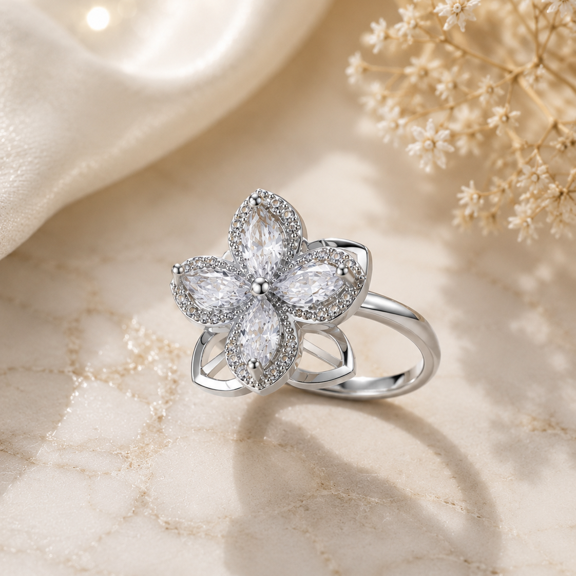 A silver flower-shaped ring with clear marquise and round stones, displayed on a beige marble surface with soft cream fabric and dried flowers in the background.